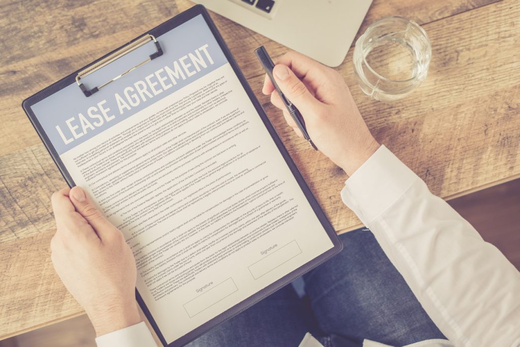 POV-photo-of-hands-holding-lease-agreement-in-clipboard-with-glass-of-water-on-table-near-laptop