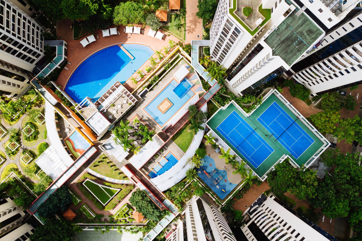 Aerial view of a residential complex with a swimming pool and tennis courts surrounded by lush greenery.